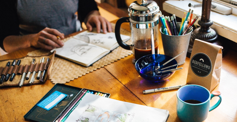 photo of art supplies splayed out on a table with a woman sketching in a notebook